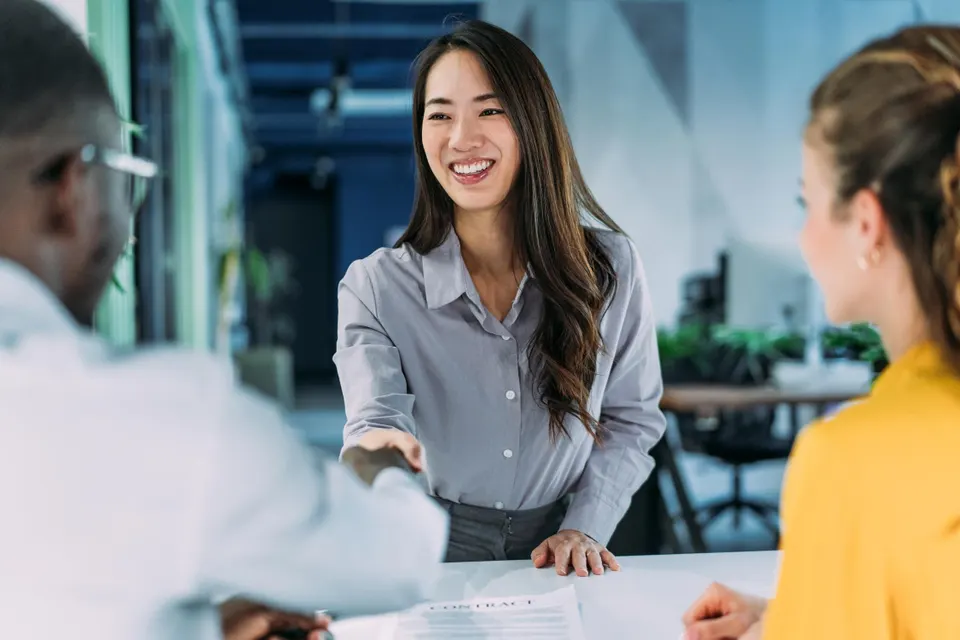 Smiling businesswoman in a gray shirt shakes hands with a colleague across a bright office conference table.