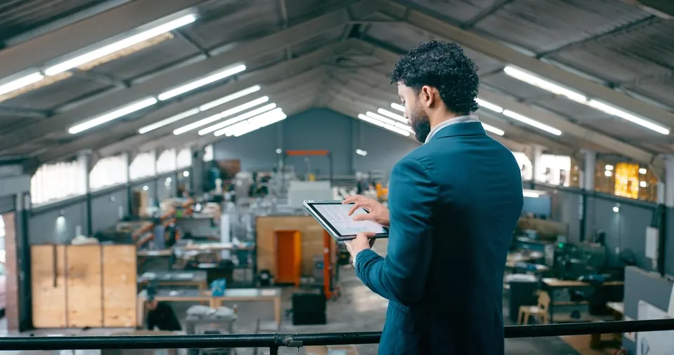 find-your-freedom Man in a blue suit stands on a balcony, using a tablet to inspect a factory floor.Industrial workspace with equipment and crates in the background.