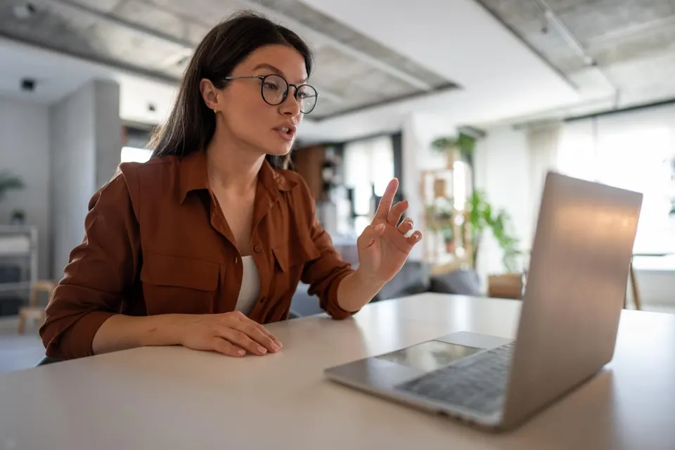 Woman with glasses sits at a white desk, gesturing with her right hand while speaking to a laptop in a bright office space.